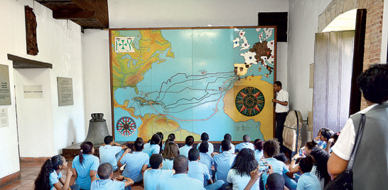 (Museo) Los estudiantes sentados y atentos escuchan las explicaciones del guía sobre las rutas utilizadas por Colón en sus viajes la isla, utilizando el mapa iluminado como instrumento didáctico. A la derecha, parte de las paredes de San Francisco.