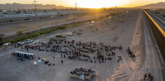 A medida que se pone el sol, los migrantes esperan afuera de una puerta en la cerca fronteriza para ingresar a El Paso, Texas, para ser procesados por la Patrulla Fronteriza, el jueves 11 de mayo de 2023.