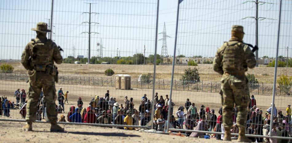 Varios migrantes esperan junto a la cerca fronteriza ante la mirada de guardias nacionales de Texas para entrar en El Paso, Texas