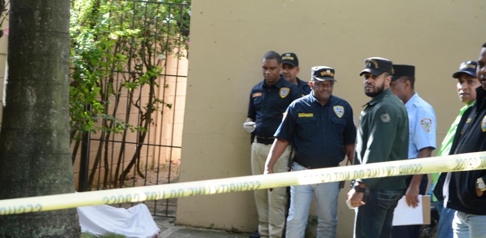 Víctima fatal. Al fondo, cubierto con un manto blanco, yace el cadáver de la asesinada sargento de la Policía, Carolina Rijo Reyes, mientras oficiales de la Direccion de Investigaciones Criminales del cuerpo policial inspeccionan la escena del crimen.