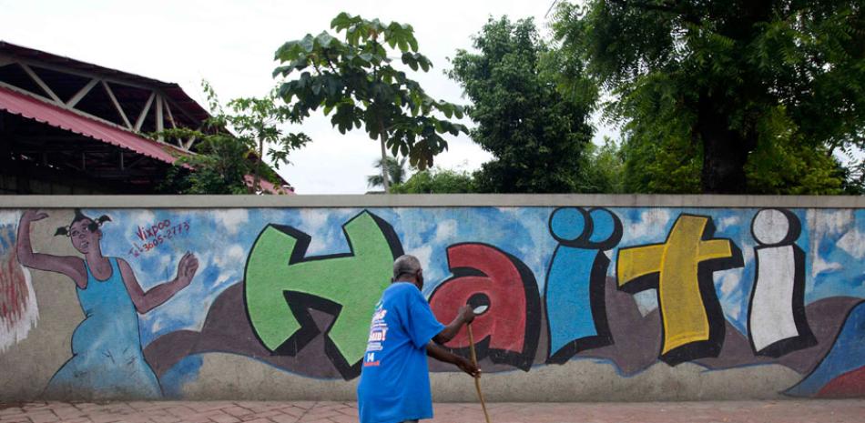 En esta imagen, tomada el 26 de mayo de 2016, un hombre barre una vereda frente a un muro de seguridad decorado con una imagen de una mujer bailando y la palabra "Haiti" escrita en colores brillantes, en Petion-Ville, un suburbio de Puerto Príncipe, Haití. AP