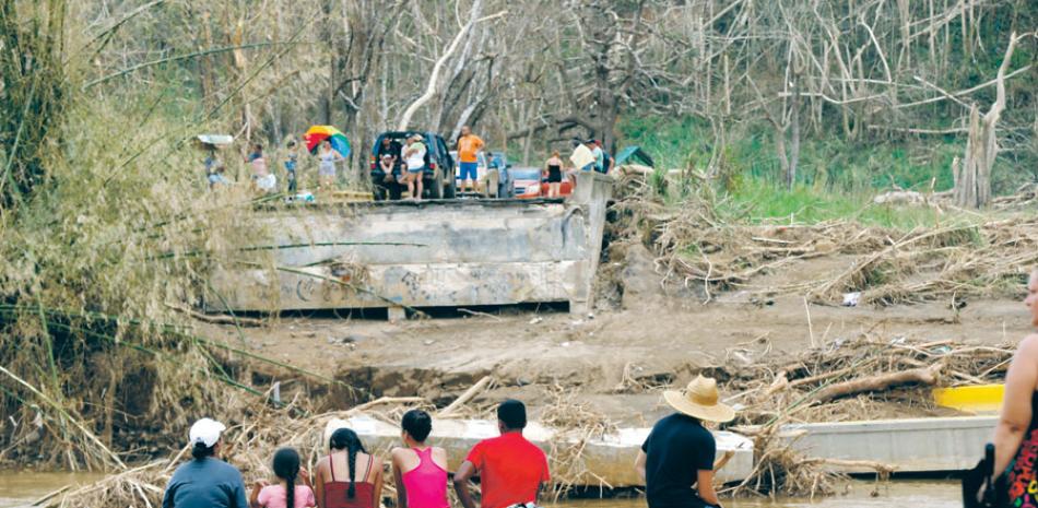 Puente. Varias personas se sientan en partes opuestas en una carretera donde había un puente que fue destruido, sobre el río Morovis, luego del paso del huracán María.