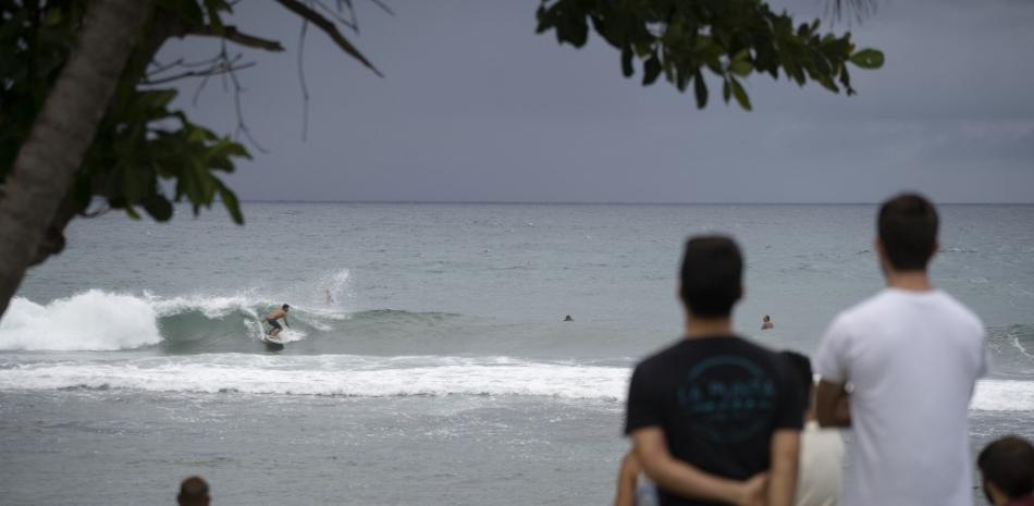 Curiosos en Puerto Rico observan el comportamiento del oleaje, mientras una persona practica surf. Fotografía de la agencia AFP.