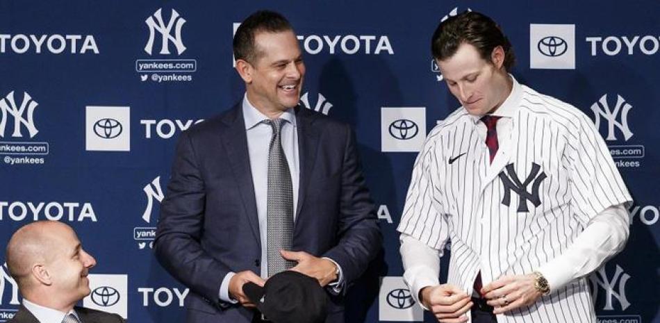 Gerrit Cole se coloca la chaqueta de los Yankees ante las sonrisas del dirigente Aaron Boone y el gerente general Brian Cashman  durante la conferencia de prensa en la que fue presentado como miembro del emblemático club.