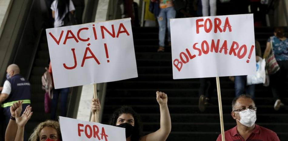 Manifestantes muestran carteles con las frases "¡Vacuna ya!" y "¡Fuera Bolsonaro" (en portugués) durante una protesta contra la gestión del presidente del país, Jair Bolsonaro, durante la pandemia del coronavirus, en una estación de autobús en Brasilia, Brasil, el 23 de diciembre de 2020. (AP Foto/Eraldo Peres)