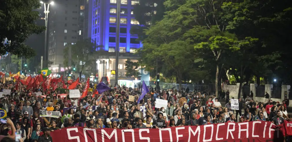 Manifestantes marchan con una bandera que dice "Somos democracia" durante una protesta para reclamar la protección de la democracia del país, en Sao Paulo, Brasil, el lunes 9 de enero de 2023. (AP Foto/Andre Penne