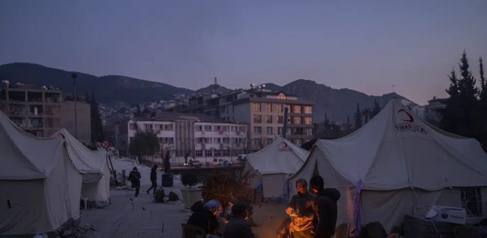 Varias personas se reúnen el lunes 13 de febrero de 2023 alrededor de una fogata en un campamento temporal para residentes que perdieron su hogar tras un sismo en Antioquía, en el sureste de Turquía. (AP Foto/Bernat Armangue)