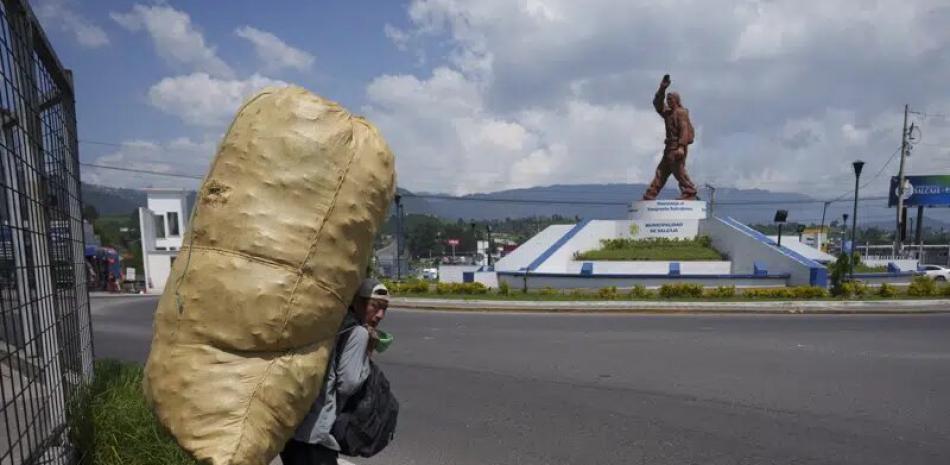 Un hombre carga con una gran bolsa de latas recicladas mientras espera a un autobús cerca de la estatua en honor a los migrantes guatemaltecos en Salcaja, Guatemala, el lunes 12 de junio de 2023.