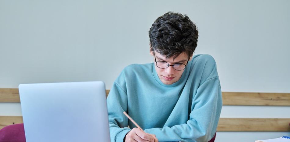 Joven estudiante universitario sentado en un escritorio en el aula, usando una computadora portátil, libros, escribiendo en un cuaderno. Educación, conocimiento, aprendizaje, cursos de idiomas, concepto juvenil