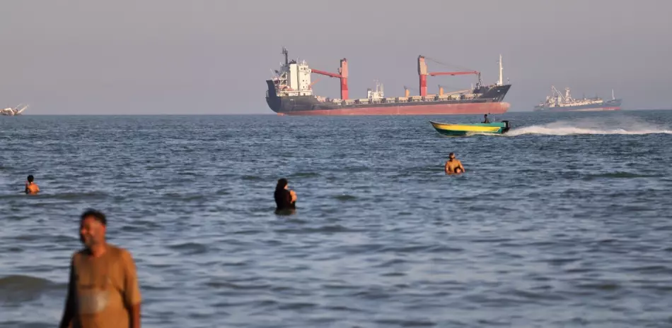 Varias personas nadan en las aguas del Estrecho de Ormuz, mientras un pequeño bote de motor pasa frente a buques de carga, el miércoles 17 de junio de 2026, cerca de Bandar Abbas, en Irán.