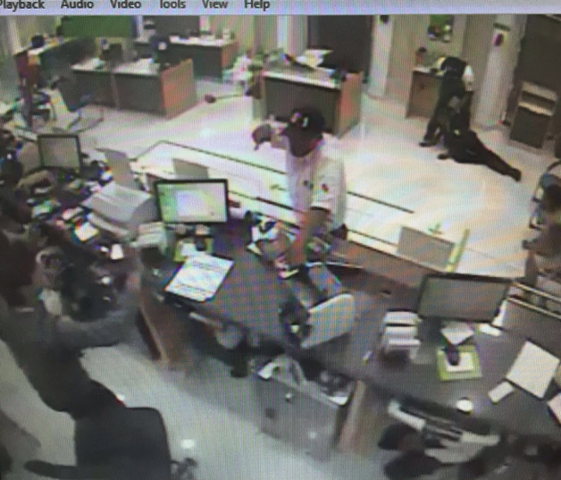Una de las escenas de la cámara de vigilancia donde se ve a uno de los asaltantes amenazando al cajero mientras el otro somete  a un hombre.