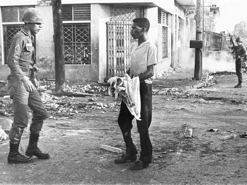 Ciudadano dominicano se topa con soldado norteamericano mientras camina por una zona de combate cargando un bebe entre sus brazos el cual enseña al militar, abril 1965.