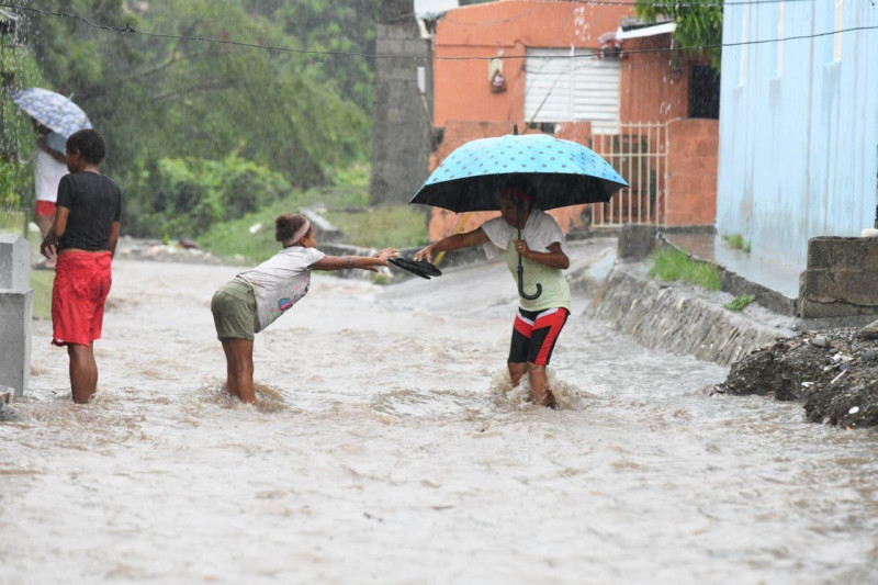 Una señora intenta pasar una cañada en Baní y es auxiliada por una niña que le toma unas chancletas. Las lluvias de la tormenta Melissa siguen causando estragos en la región sur del país