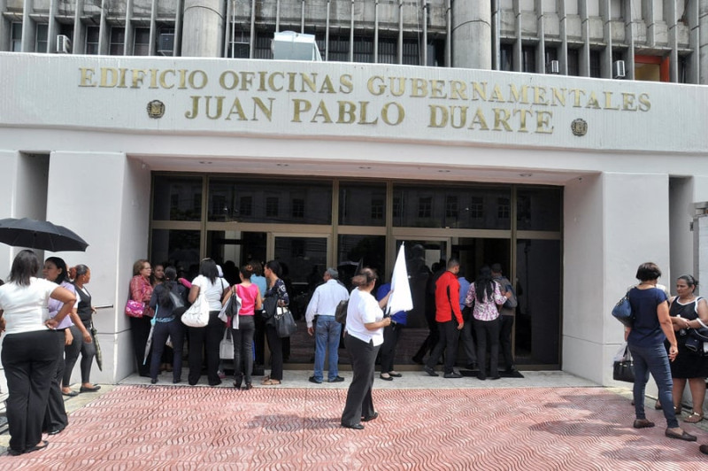 Entrada principal del Edificio de Oficinas Gubernamentales Juan Pablo Duarte, en la avenida México. En la foto se ven algunos empleados entrando al edificio y otros hablan.