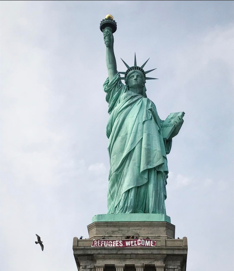 En esta fotografía, facilitada por Vitória Londero, se ve el cartel gigante con el mensaje "Refugees Welcome" (Bienvenidos refugiados, en español) colgando en el pedestal de la Estatua de la Libertad en Nueva York. (Vitória Londero via AP)