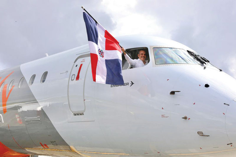 Abinader levanta la bandera nacional a través de una ventanilla de piloto de avión de la empresa Embraer.