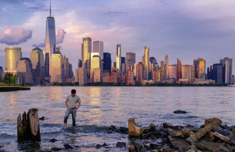 Un hombre comprueba dónde pisa mientras camina por el agua en el Canal Morris en Jersey, Nueva Jersey, mientras se pone el sol con Manhattan de fondo