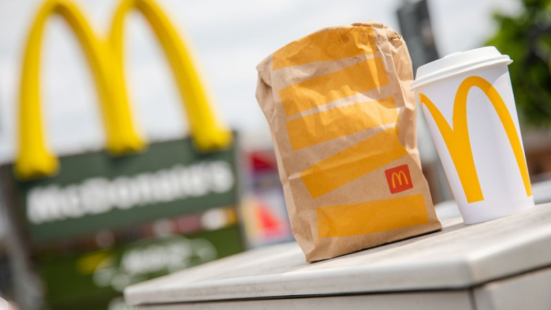 Una bolsa para llevar de McDonald's y un puesto de vasos para llevar que la acompaña frente a una sucursal de McDonald's.