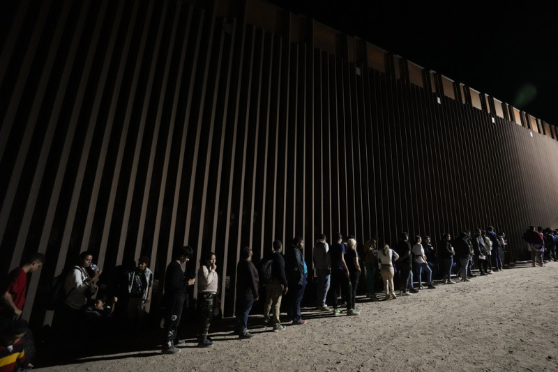 Un grupo de personas hacen fila junto al muro fronterizo mientras esperan para solicitar asilo