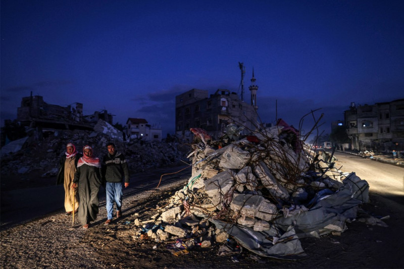 Vehículos iluminan una carretera mientras unos hombres pasan junto a los escombros de un edificio destruido por los bombardeos israelíes en Rafah