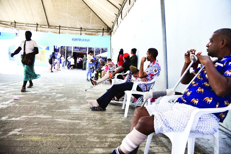 Sentados al lado de una pared, pacientes esperan sus turnos, para gestiones de asistencia médica, en el área externa de hospital en la capital.
