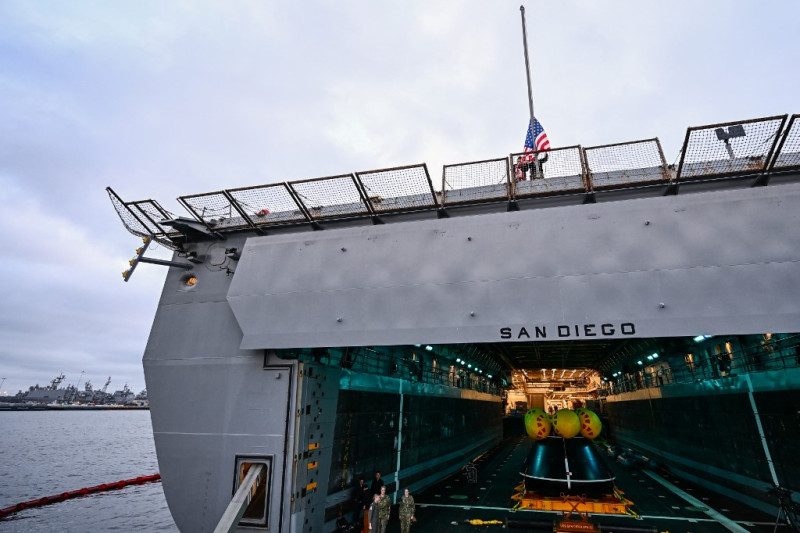 El personal militar se mantiene firme mientras la bandera estadounidense se baja al atardecer desde la popa mientras la cápsula de prueba del módulo de tripulación Orion se asienta en la cubierta del pozo a bordo del USS San Diego (LPD-22) en la Base Naval de San Diego después de la prueba de recuperación en marcha 11 en San Diego, California, el 28 de febrero de 2024.