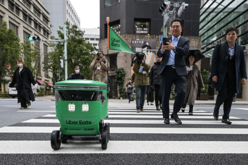 Miembros de los medios de comunicación y transeúntes caminan junto a un robot no tripulado mientras cruza la calle durante una demostración de un servicio de entrega de robots de Uber Eats Japan, Mitsubishi Electric y el desarrollador de robots Cartken en el centro de Tokio el 5 de marzo de 2024.