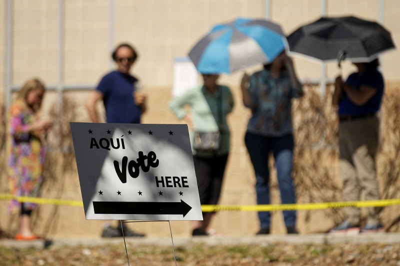 Votantes usan sombrillas para cubrirse del sol mientras esperan formados en un centro de votación, el martes 5 de marzo de 2024, en San Antonio.