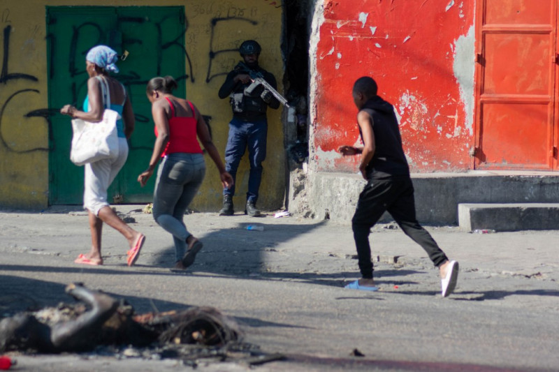 Varias personas caminan junto a cadáveres quemados y un agente de policía armado que vigila una calle tras otra jornada violencia de bandas el 21 de marzo de 2024, en Puerto Príncipe, Haití