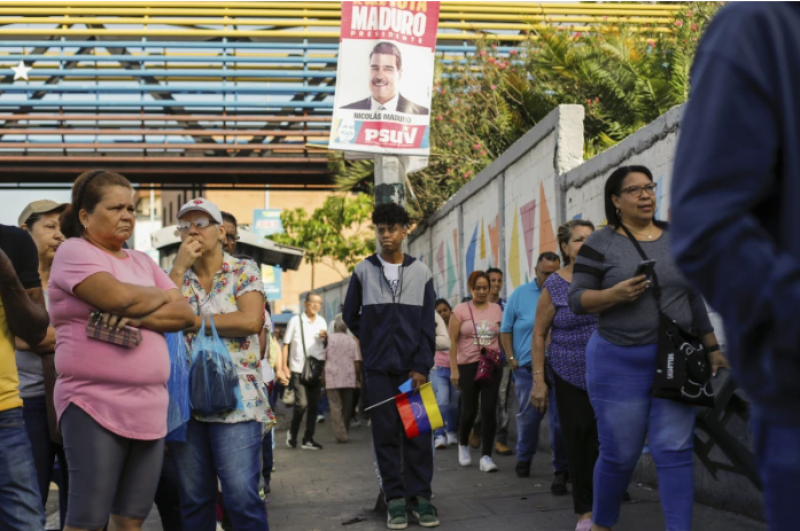 Los votantes hacen fila en un colegio electoral donde cuelga un cartel de campaña del presidente Nicolás Maduro durante las elecciones presidenciales en Caracas, Venezuela, el domingo 28 de julio de 2024. (AP Foto/Cristian Hernández)