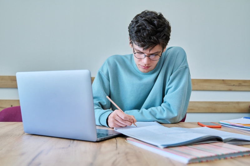 Joven estudiante universitario sentado en un escritorio en el aula, usando una computadora portátil, libros, escribiendo en un cuaderno. Educación, conocimiento, aprendizaje, cursos de idiomas, concepto juvenil