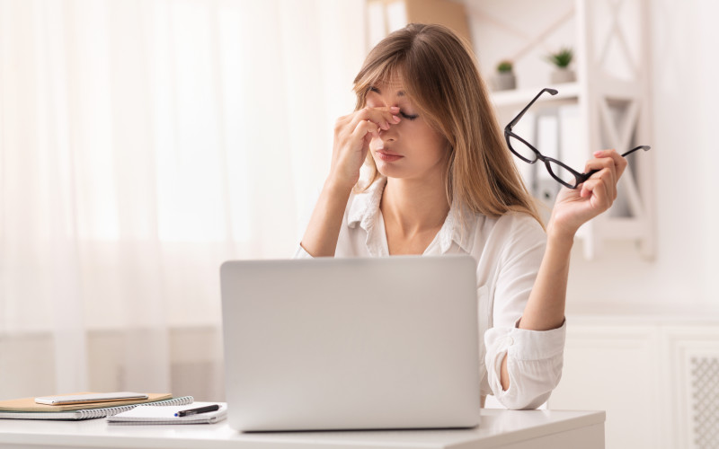 Fatiga de los ojos. Trabajadora masajeándose el puente nasal al sentir hipertensión ocular sentada ante su portátil en el lugar de trabajo. Foto: Prostock-studio - stock.adobe.co