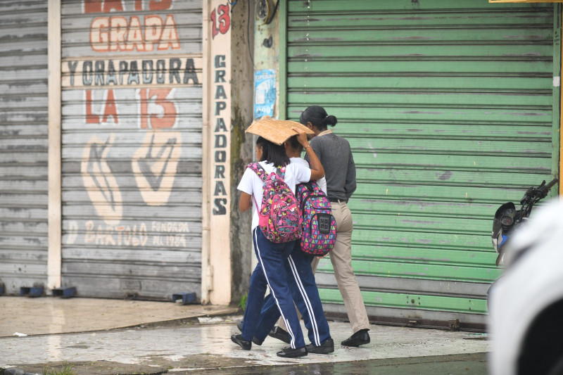 Dos estudiantes se cubren de la lluvia con un carton mientras caminan en una acera del Distrito Nacional.