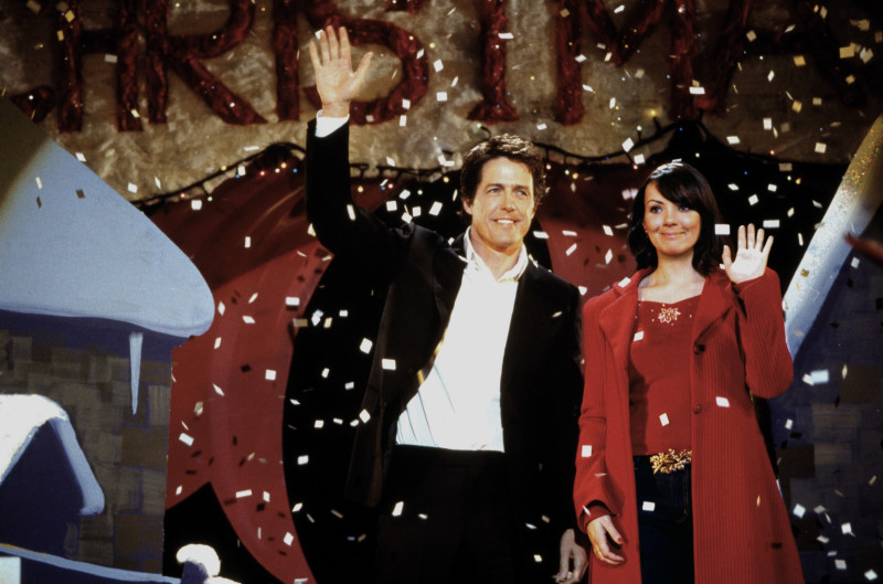 En "Love Actually" (2003), varios personajes se entrelazan durante la función navideña de un colegio, un momento cargado de sorpresas y revelaciones. Entre ellos, el primer ministro (Hugh Grant) y su secretaria Natalie (Martine McCutcheon) ven su romance destapado ante la atónita audiencia, en una escena que combina humor y ternura, destacando las conexiones inesperadas que las fiestas pueden traer. ©Universal/Courtesy Everett Collection.  ©Universal/Courtesy Everett Collection