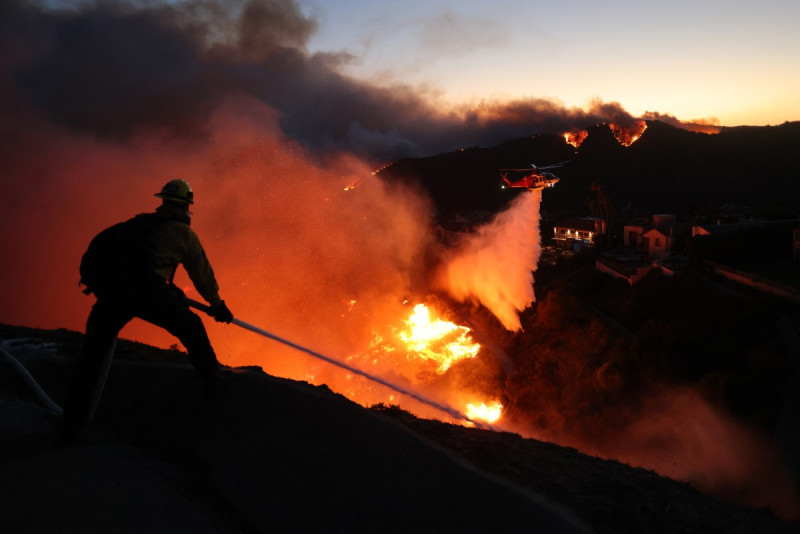 Un bombero apaga las llamas mientras un helicóptero arroja agua alrededor de las casas amenazadas por el incendio Palisades impulsado por el viento en Pacific Palisades, California, 7 de enero de 2025.