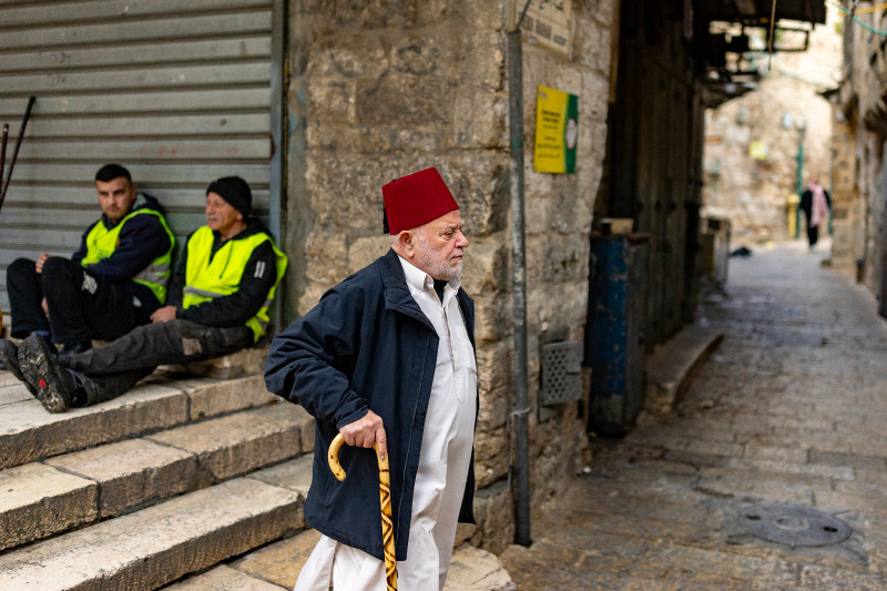 Un anciano palestino pasa junto a barrenderos que están en su descanso en la Ciudad Vieja de Jerusalén el 23 de marzo de 2026.