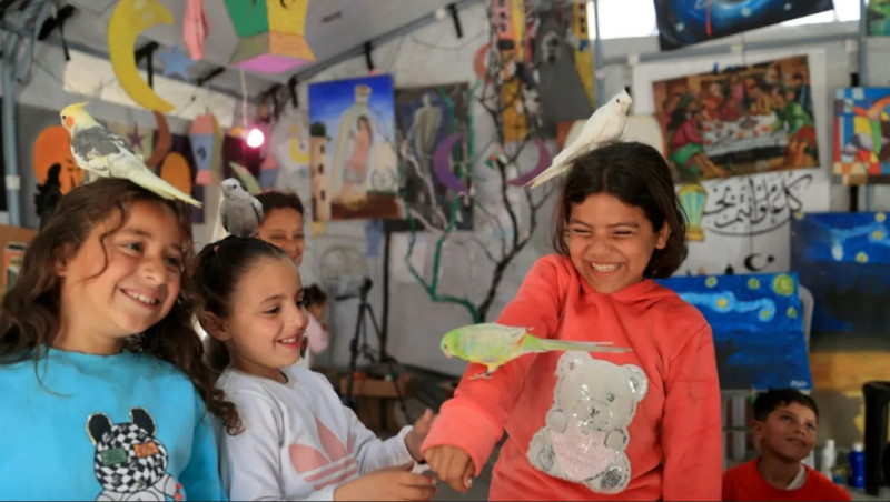 Niñas de Gaza sonríen al interactuar con unos pájaros se les posan en la cabeza y en el brazo.