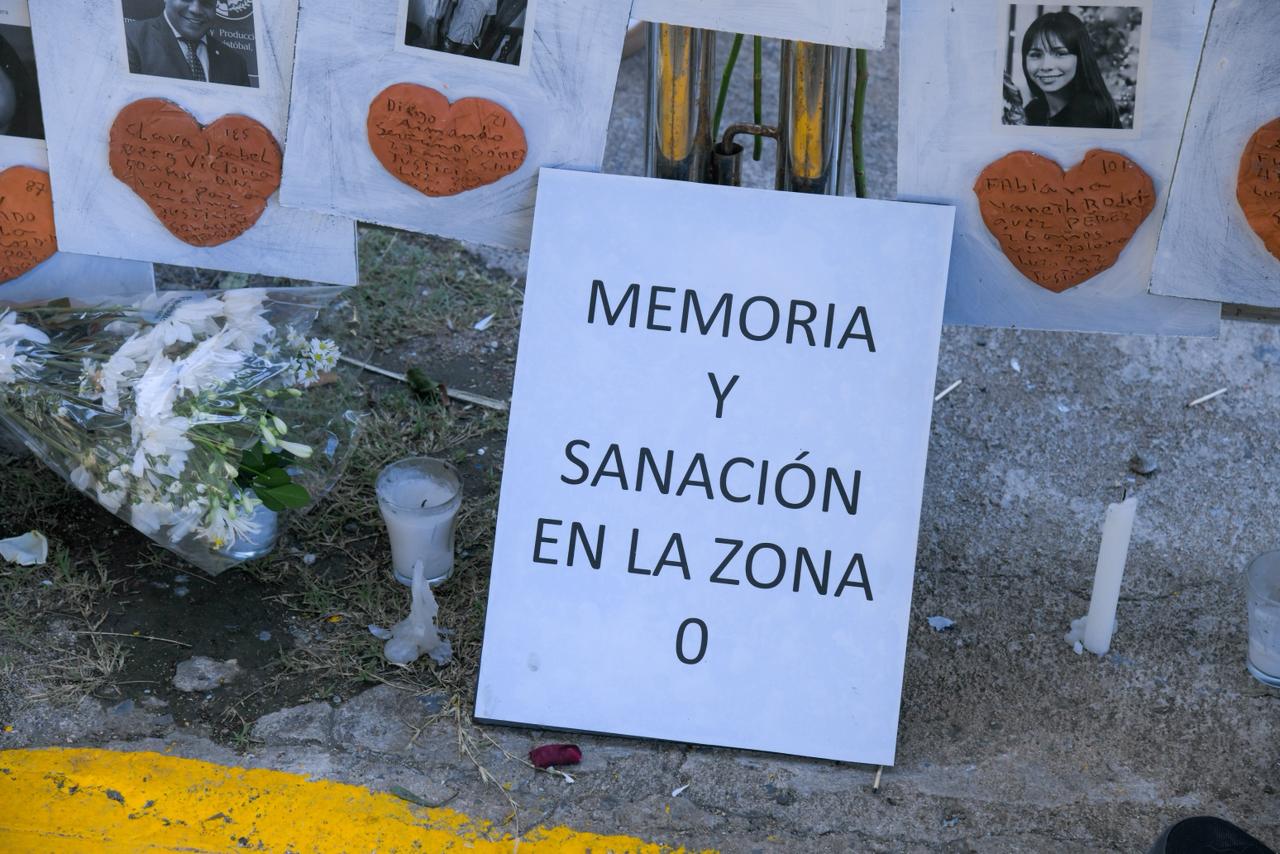 Cartel con el mensaje “Memoria y sanación en la Zona 0” acompaña flores y velones en honor a las víctimas.